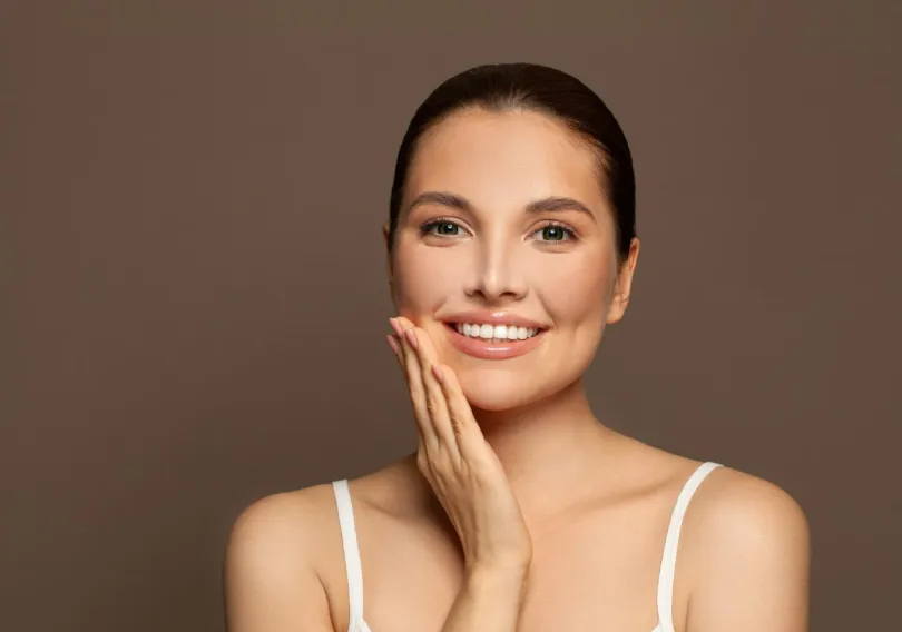 Smiling woman with clear skin touching her cheek against a brown background.