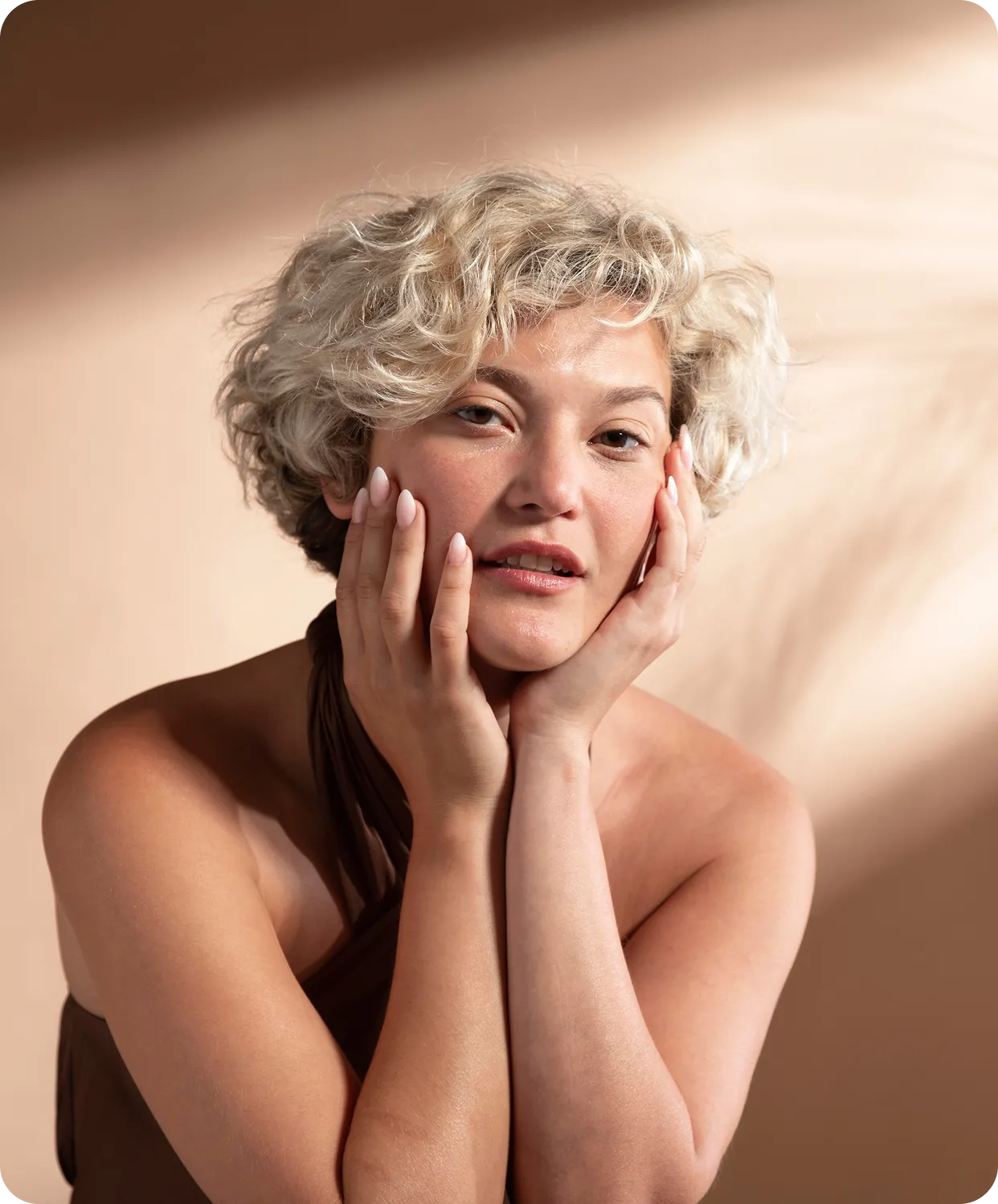 Blonde woman with curly hair gently touching her face, wearing a brown halter top against a beige background.