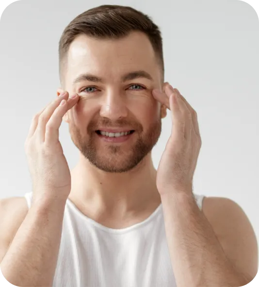 Smiling man in a white tank top touching his face with both hands against plain background.