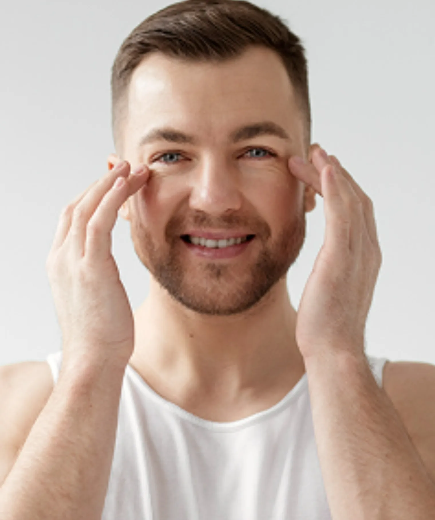 Smiling man with beard touching face skin with fingers wearing white sleeveless top.