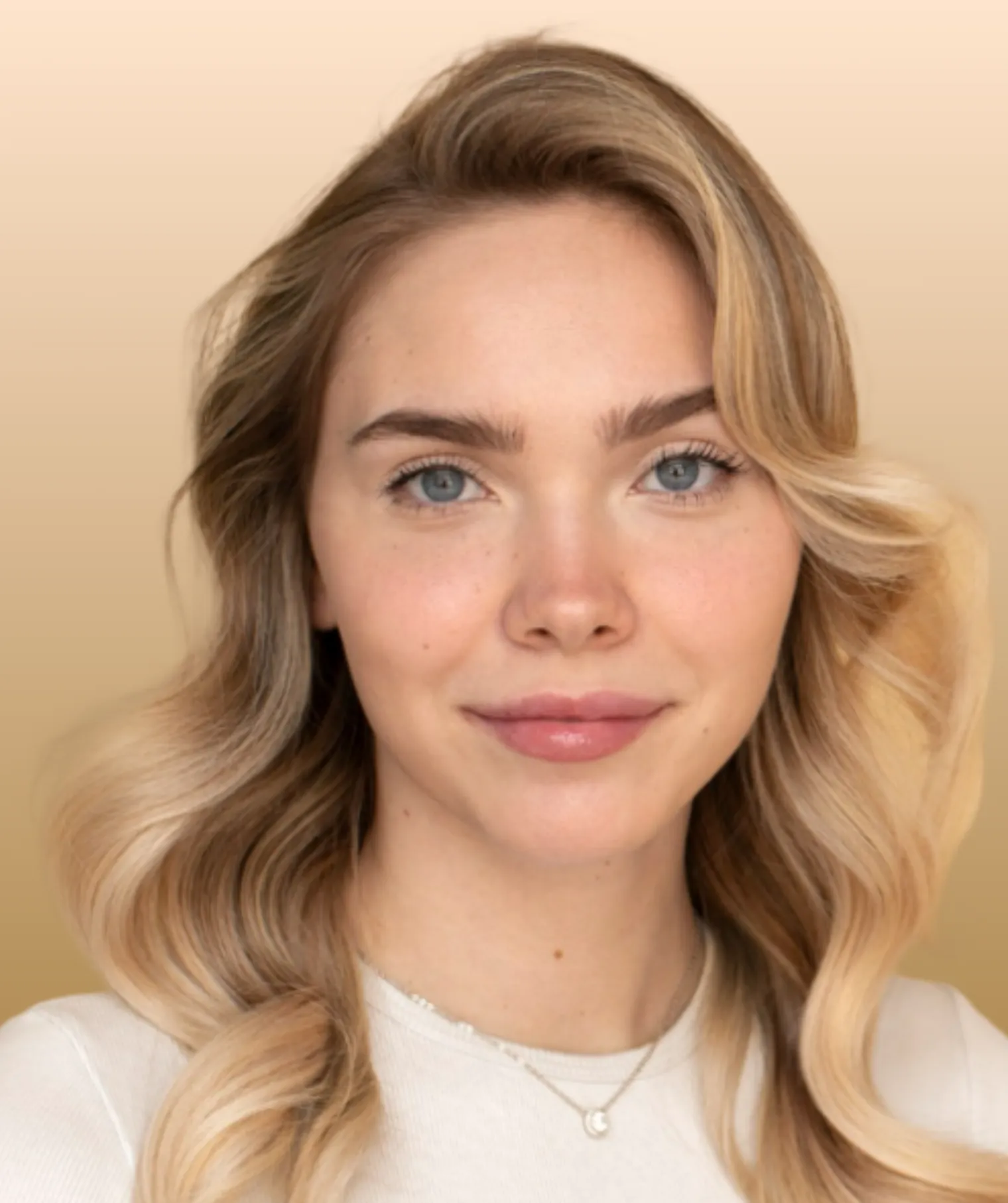 Portrait of a smiling young woman with blonde wavy hair and blue eyes wearing a white top and necklace.