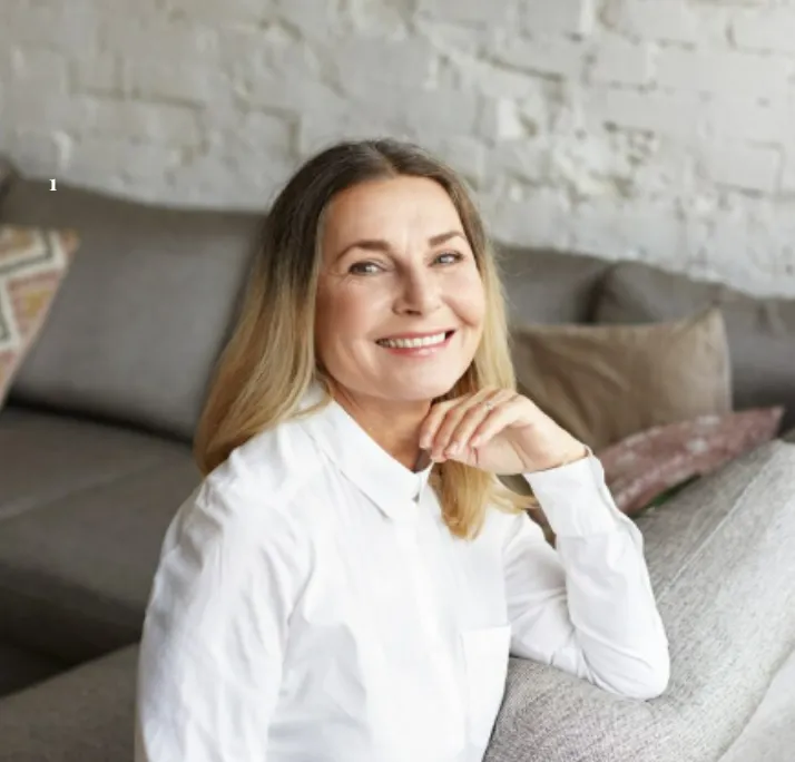 Smiling middle-aged woman with light hair leaning on a couch in a cozy living room.
