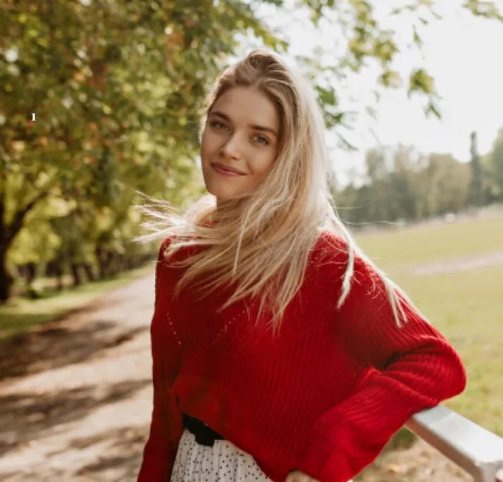 Smiling young woman with long blonde hair wearing a red sweater outdoors near a park fence.