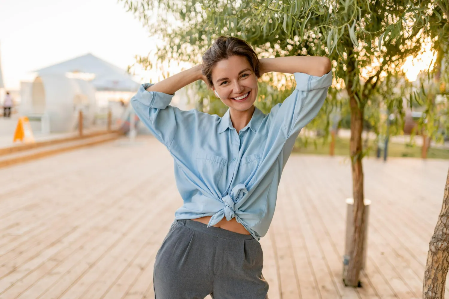 Smiling woman in blue shirt and gray pants posing with hands behind head outdoors.