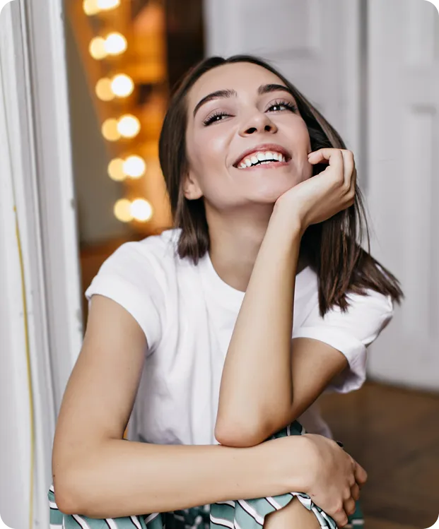 Smiling young woman in white shirt sitting indoors with lights blurred in the background