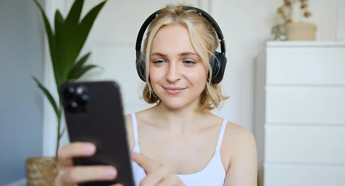 Smiling blonde woman wearing headphones using smartphone in a bright room with plant and dresser