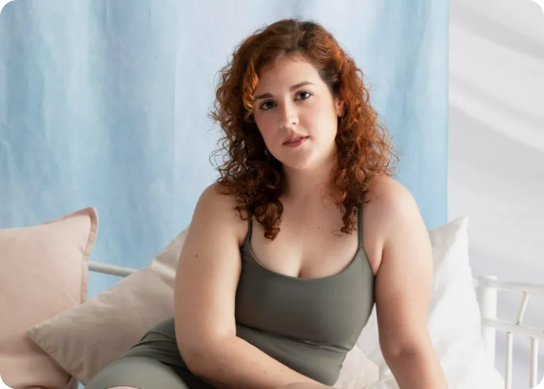 Woman with curly red hair wearing a green tank top sitting on a bed with pillows and light blue backdrop.