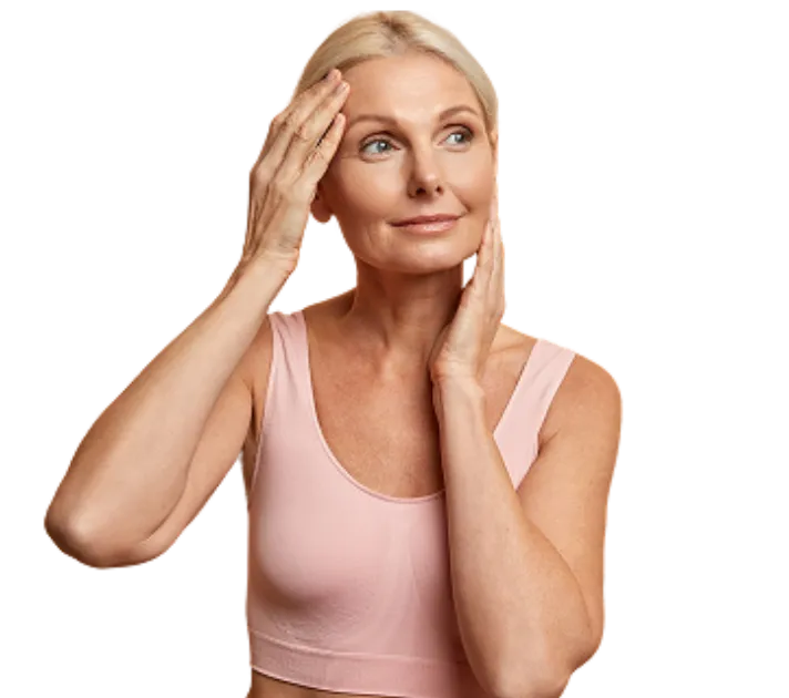 Smiling older woman in pink top touching her face and forehead, looking thoughtful.