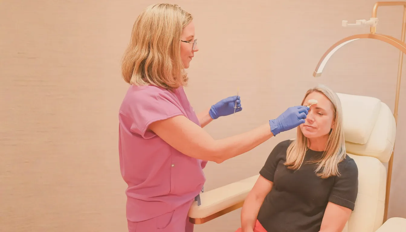 A healthcare professional wearing gloves applies a cosmetic treatment to a seated patient's forehead.