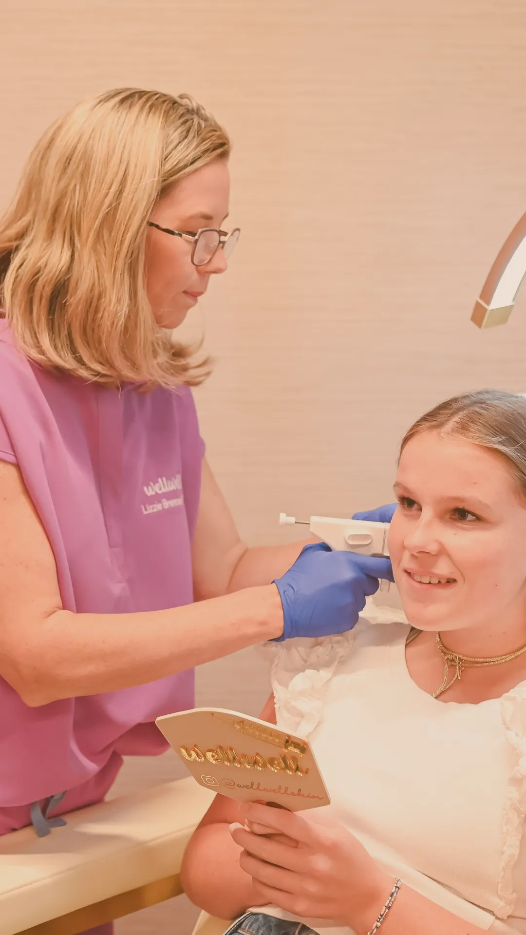 Woman wearing purple examines a girl's ear with a medical device as the girl holds a mirror and smiles.