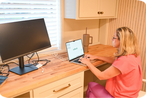 Woman with glasses typing on a laptop at a kitchen counter near a monitor and cup.
