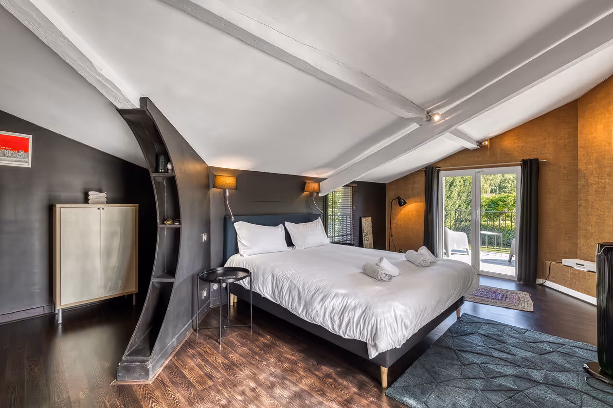 Chambre avec grand lit blanc, murs sombres, lampe murale, et porte fenêtre donnant sur balcon vert.