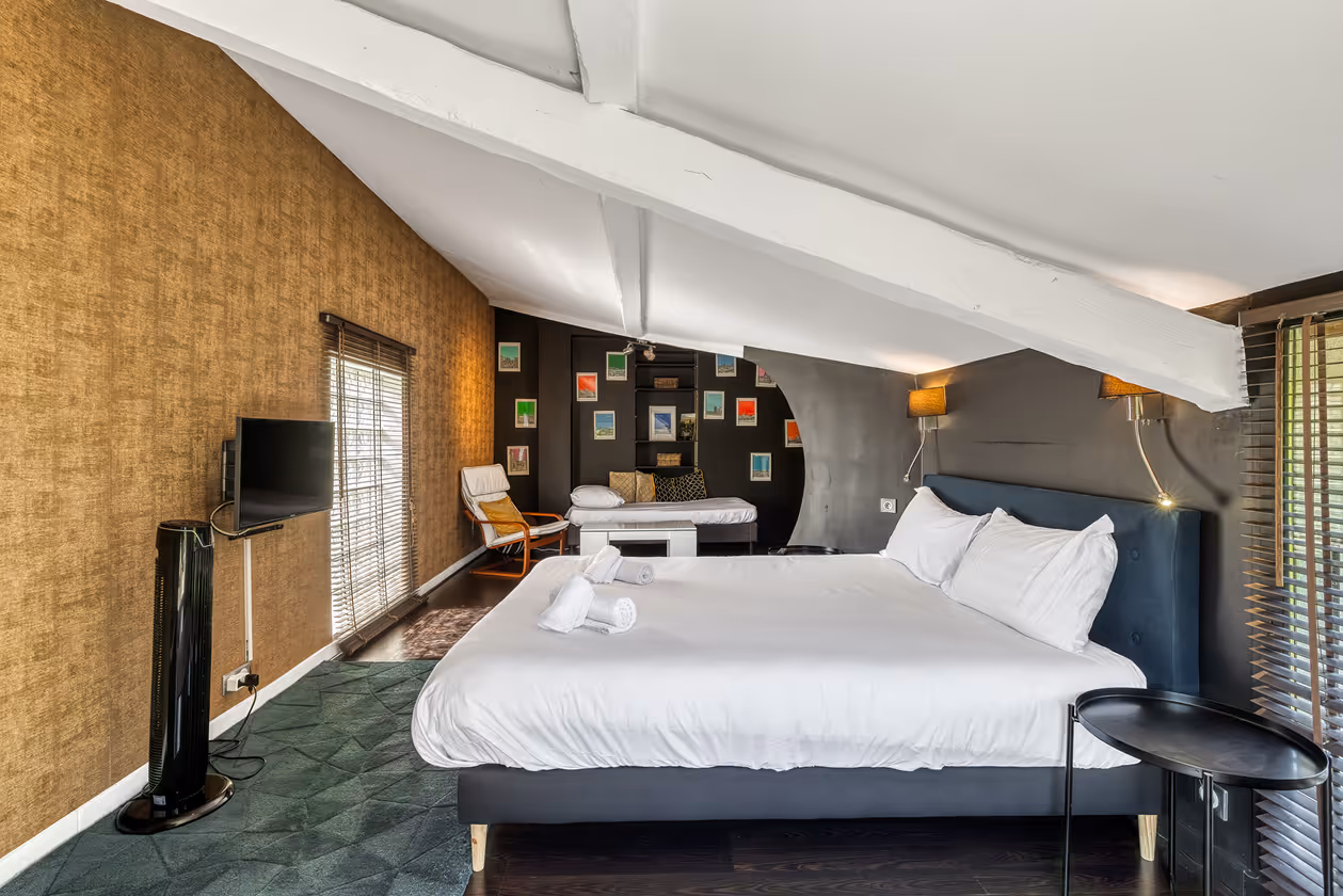 Chambre avec grand lit blanc, mur beige, fauteuil, table basse, TV et poutres blanches au plafond.