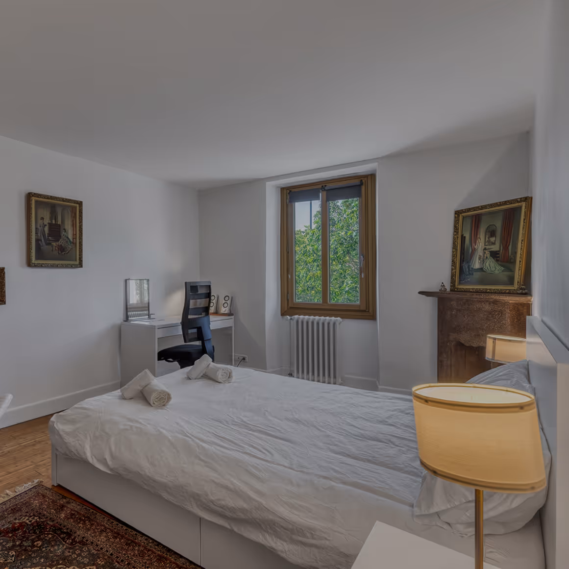 Chambre avec lit double, bureau, chaise, tapis, lampe, deux tableaux, et fenêtre sur arbres verts.