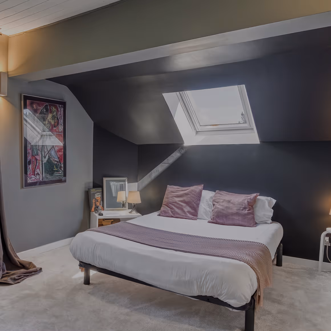 Chambre avec mur et plafond noirs, lit blanc, coussins et jeté mauve, fenêtre de toit et lampes.