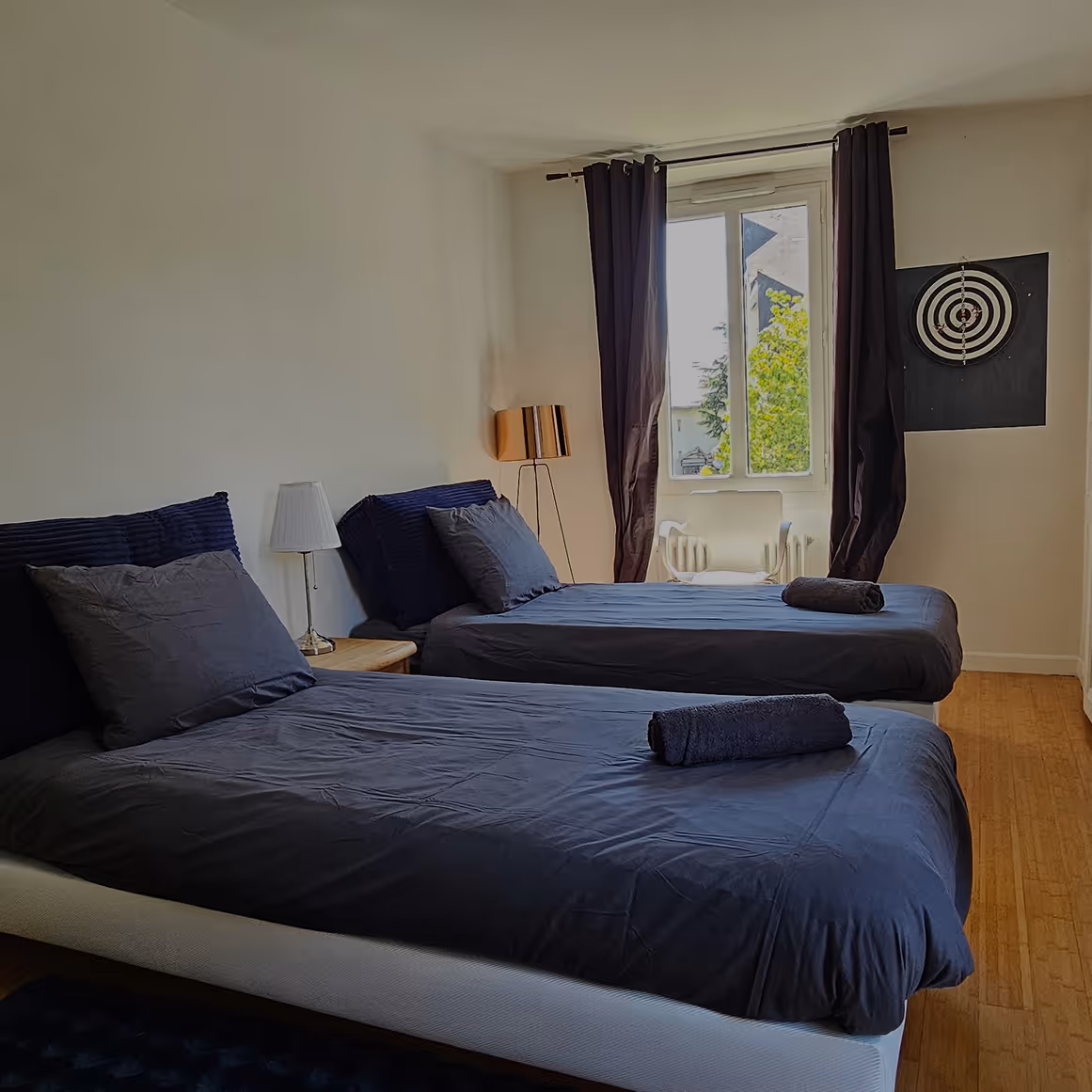 Chambre avec deux lits simples aux draps bleus, deux lampes, rideaux foncés et une cible au mur.