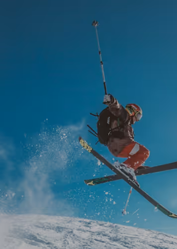 Skier in a red outfit performing a jump with poles and crossed skis on snow under a blue sky.