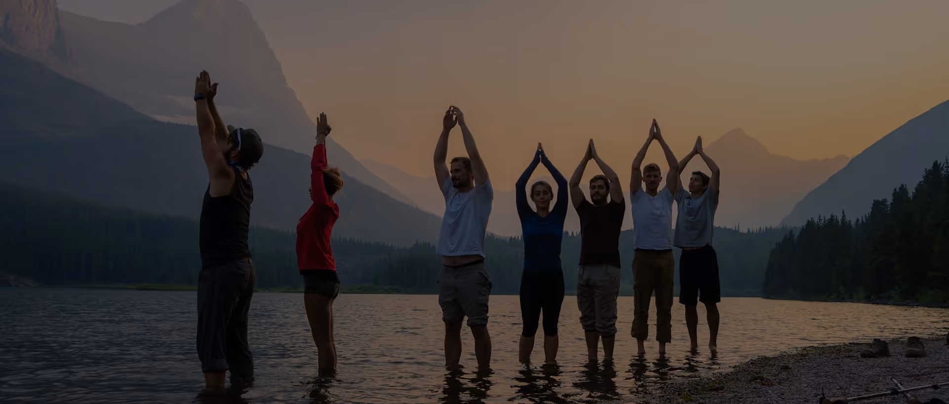 Un groupe de personnes debout dans l'eau au coucher du soleil avec des bras levés en position de yoga.