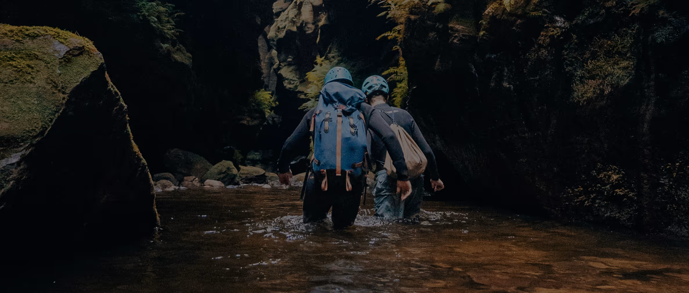 Deux personnes en tenue de randonnée traversent une rivière entre des falaises rocheuses ombragées.