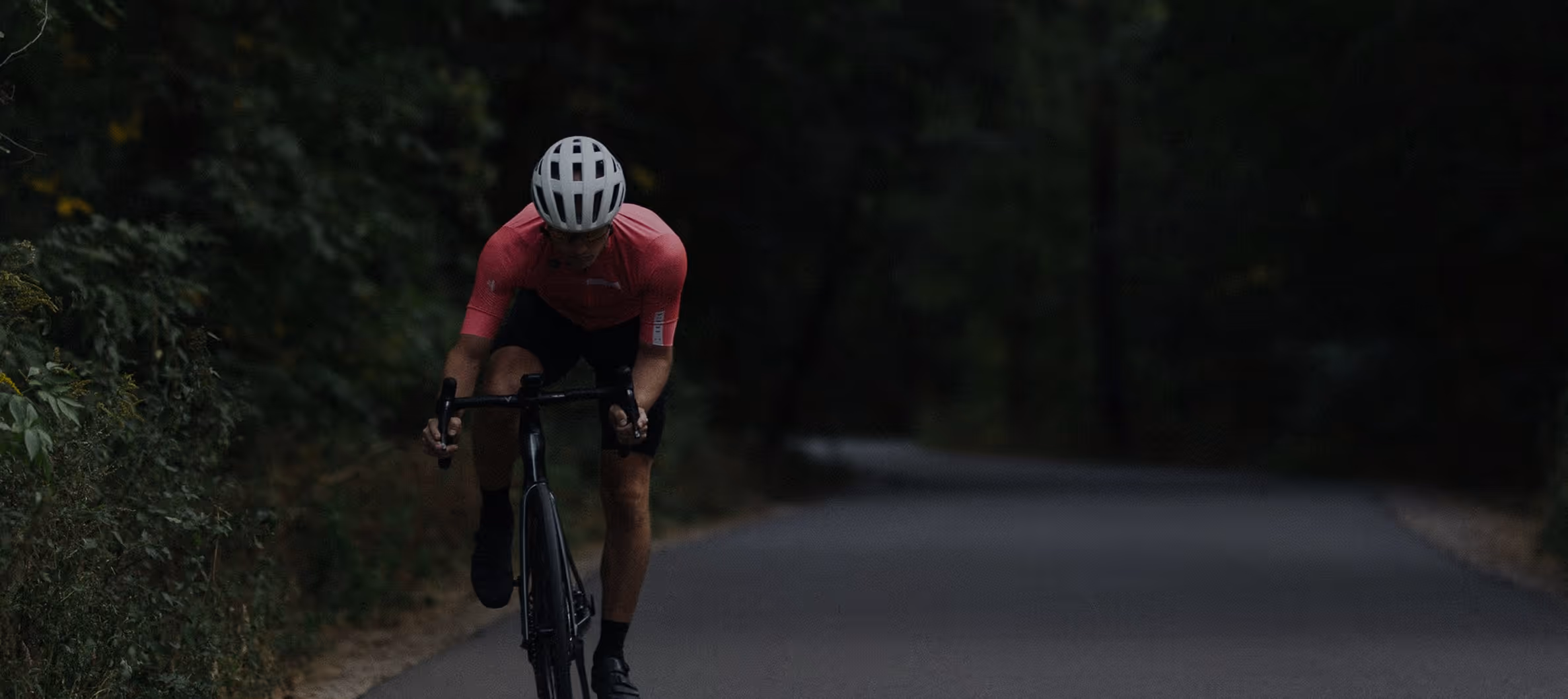 Cycliste en maillot rose et casque blanc sur une route entourée d'arbres sombres.