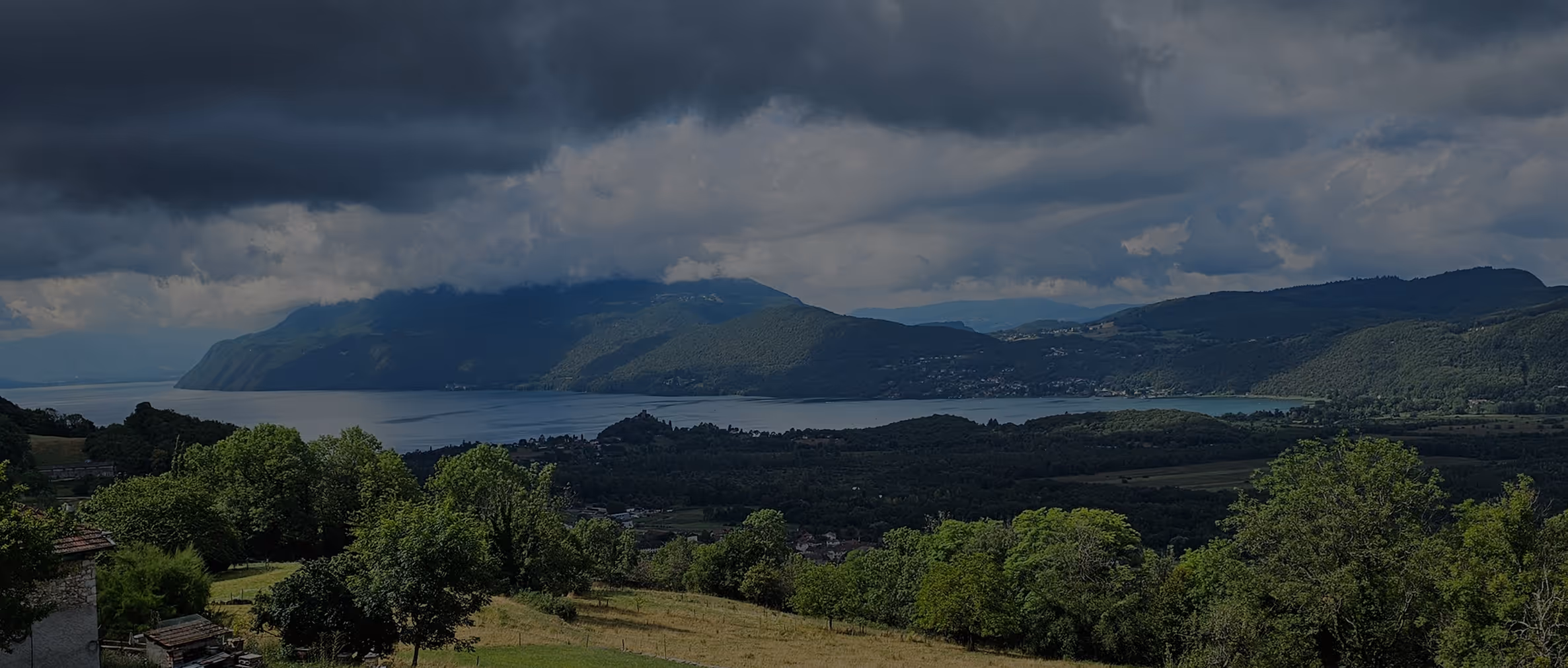 Vue panoramique sur un lac entouré de montagnes sous un ciel nuageux et arbres au premier plan.