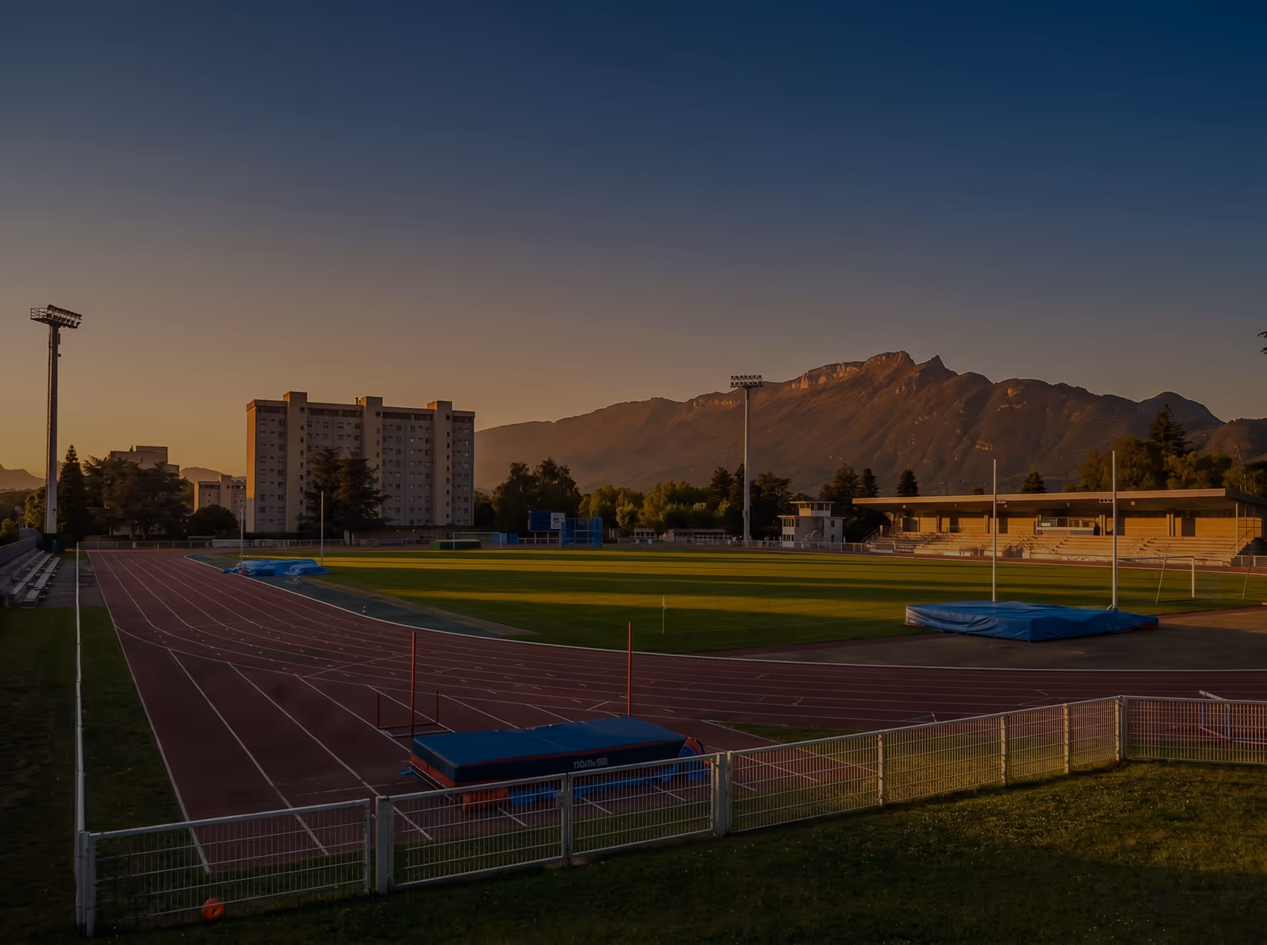 Piste d'athlétisme et terrain de sport au crépuscule avec montagnes en arrière-plan.