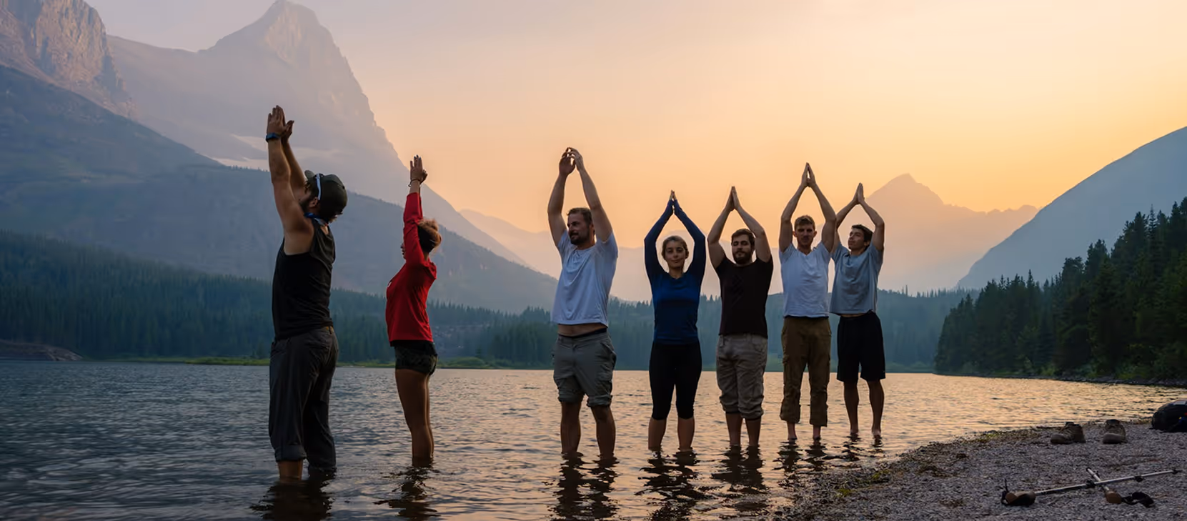 A group of seven people standing in a lake doing yoga at sunset in the mountains.