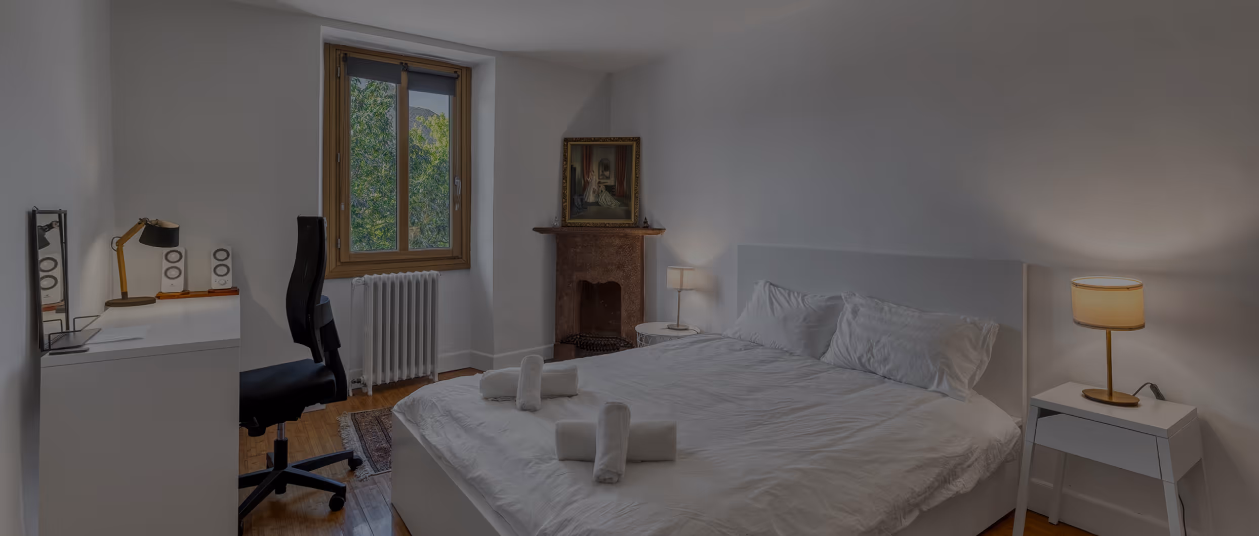 Chambre simple avec lit double, linge blanc plié, bureau noir, deux lampes et tableau au mur.