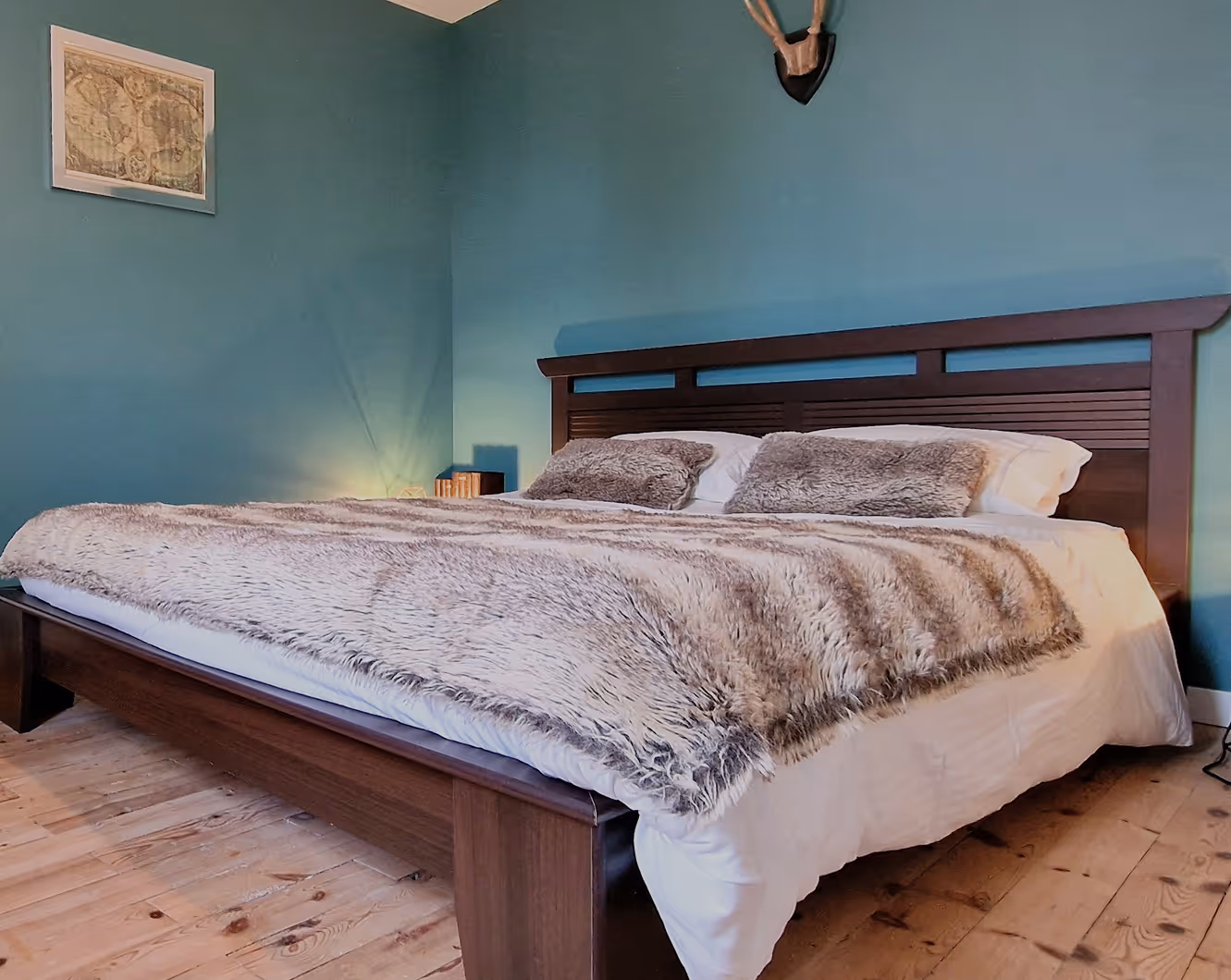 Chambre avec grand lit en bois, linge blanc et couverture moelleuse grise, murs bleus et plancher en bois.