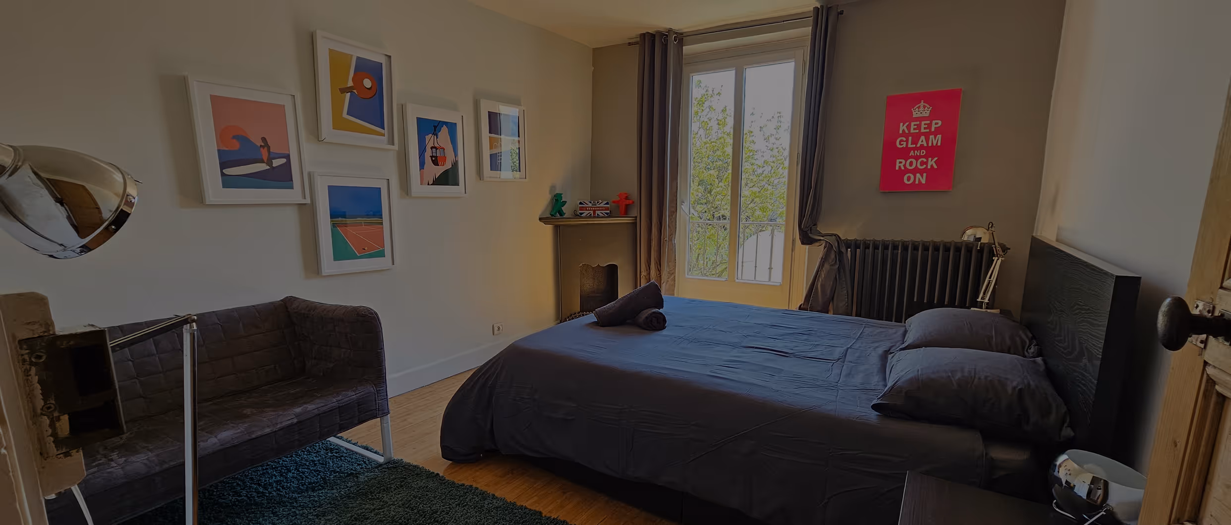 Room with a double bed draped with grey linen, a window, curtains, a radiator, and a pink 'Keep glam and rock on' poster.