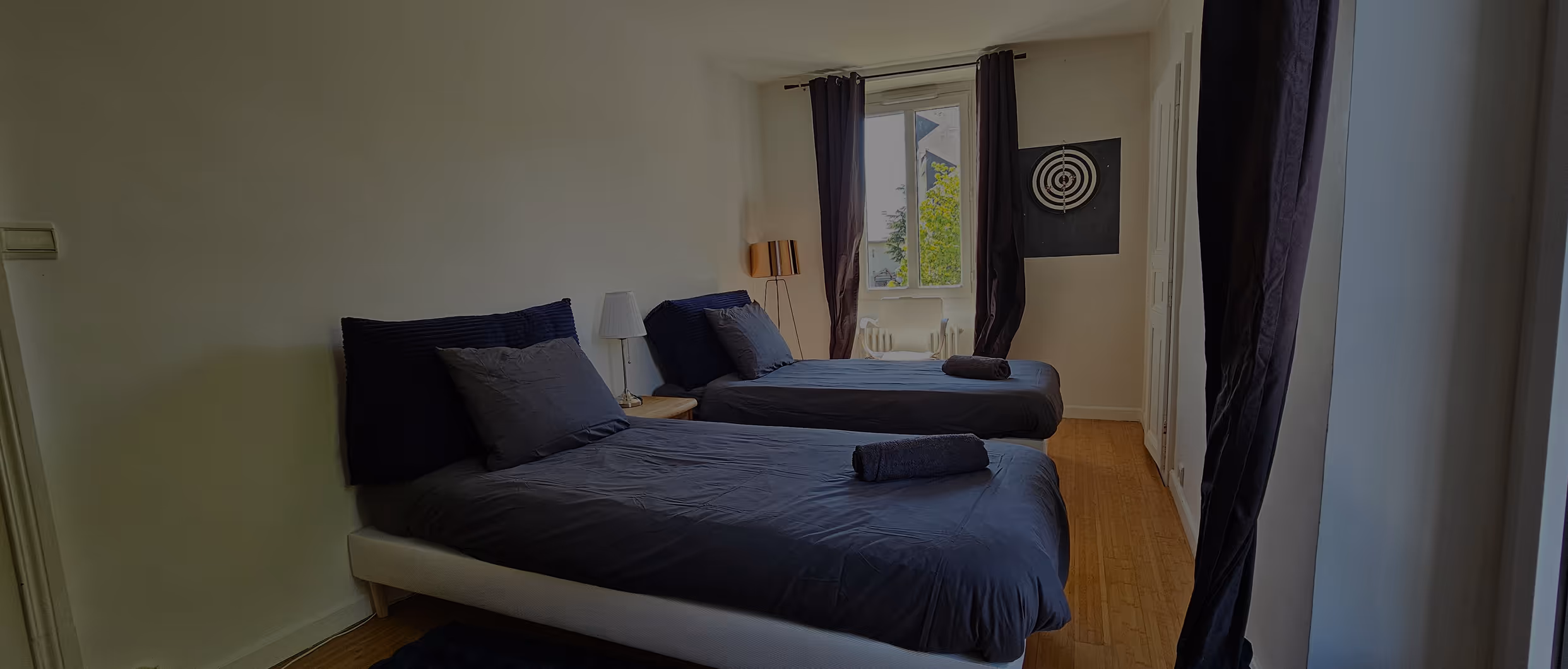 Chambre avec deux lits simples à draps et coussins gris, fenêtre avec rideaux noirs et un jeu de fléchettes.