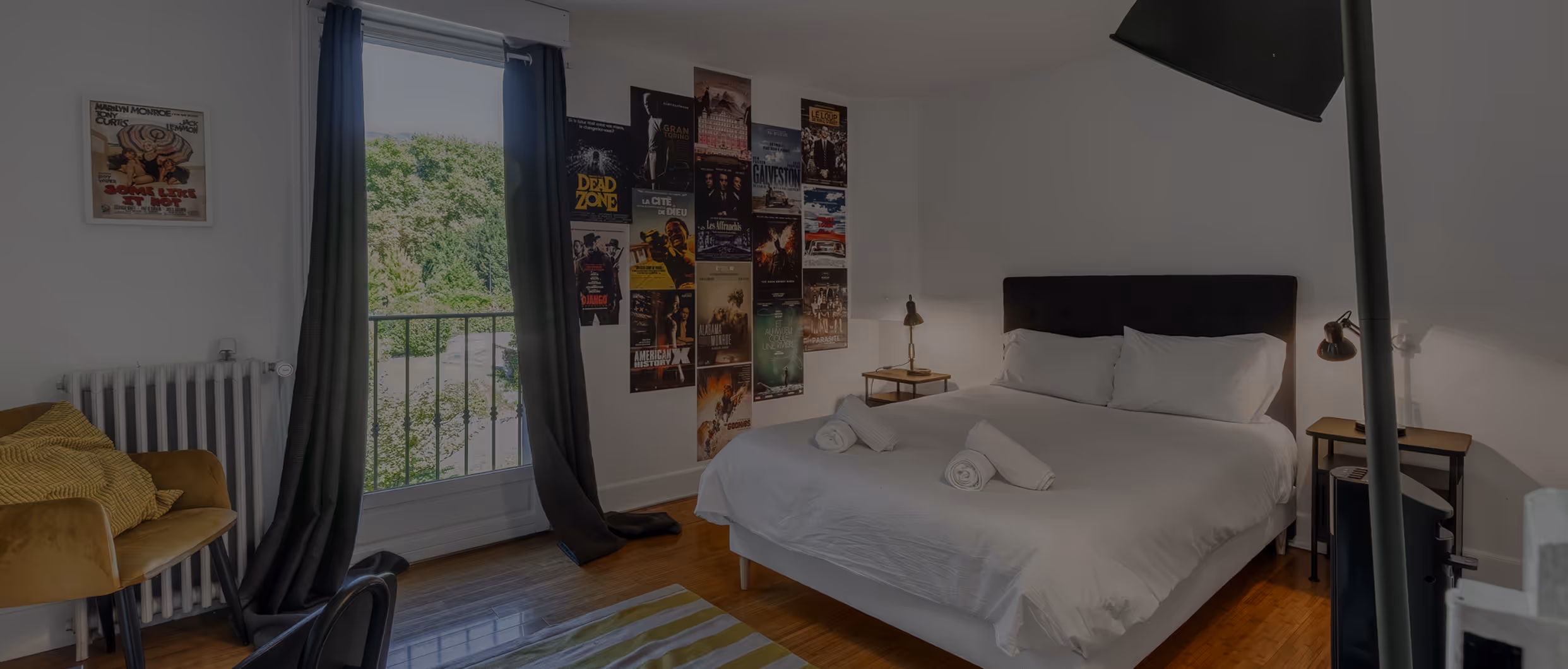 Chambre avec lit double blanc, murs avec affiches, fauteuil jaune et grande fenêtre avec rideaux noirs.
