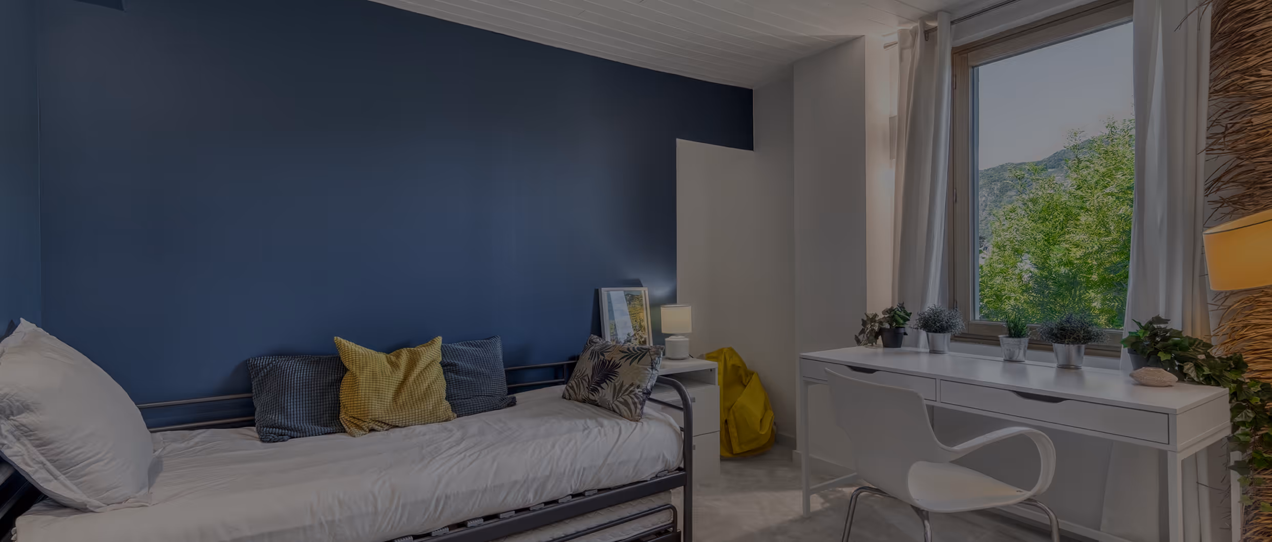Chambre avec lit simple, coussins colorés, bureau blanc, chaise, et vue sur arbres par la fenêtre.
