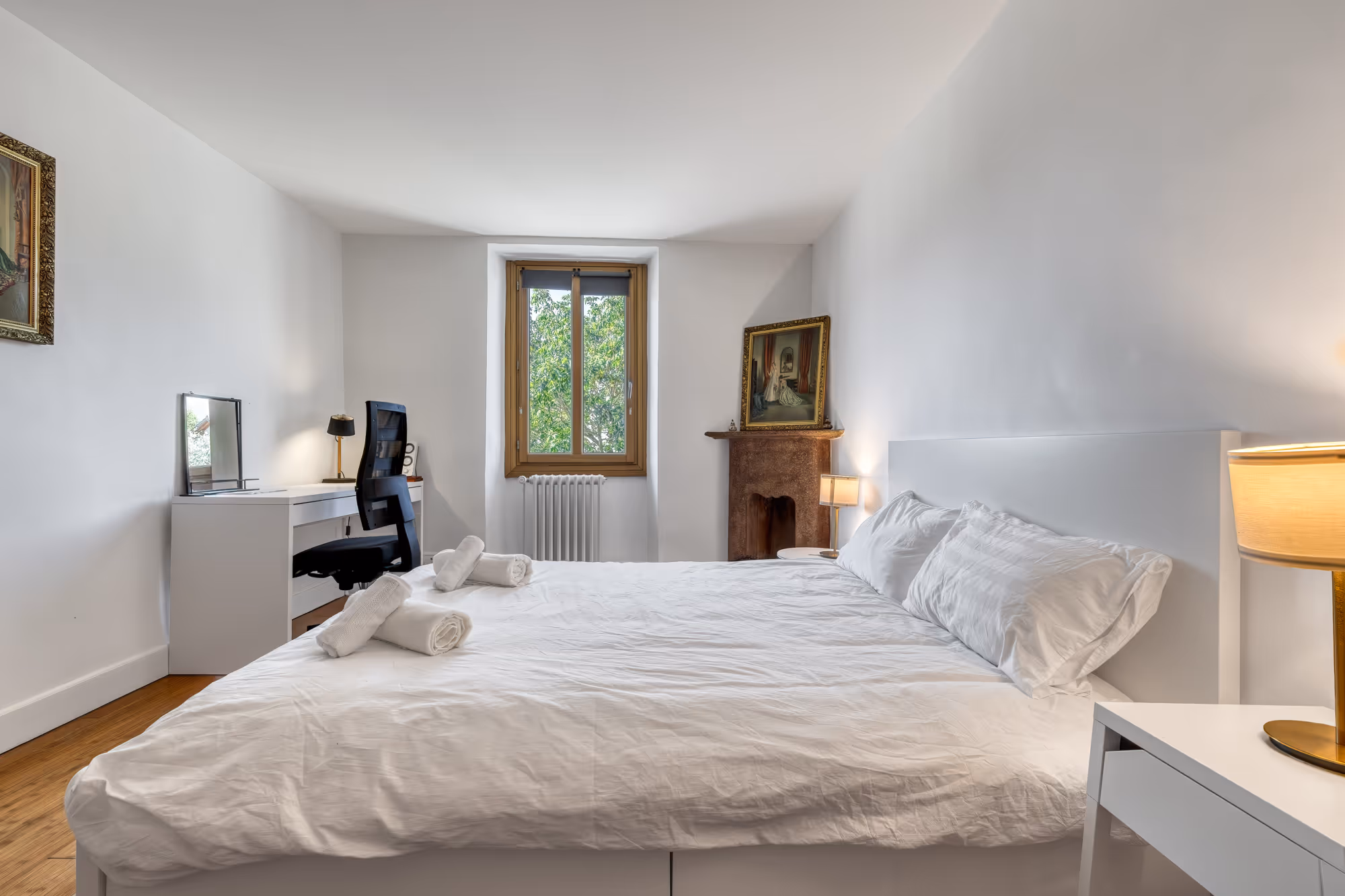 Chambre moderne avec lit double, chaise, canapé, murs décorés, et grande fenêtre avec stores.