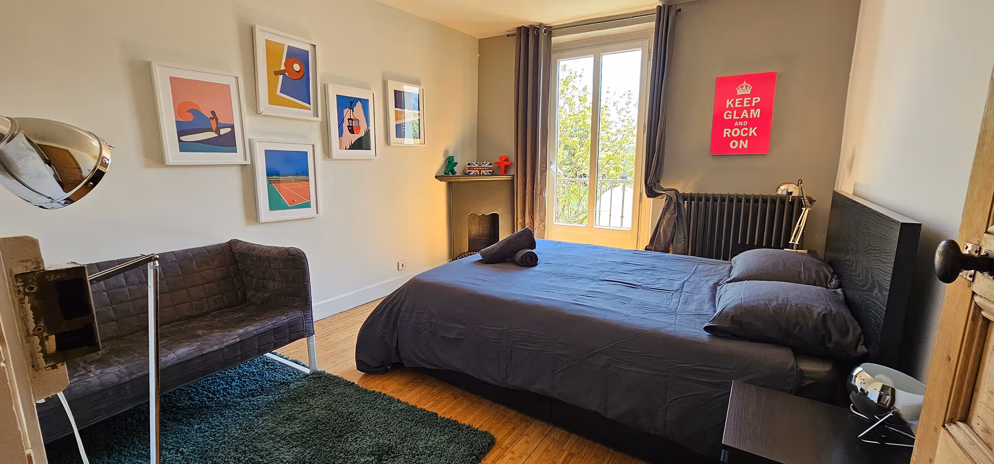 Room with a grey double bed, sofa, green rug, five colorful paintings, a window, and a pink 'Keep Glam and Rock On' poster.