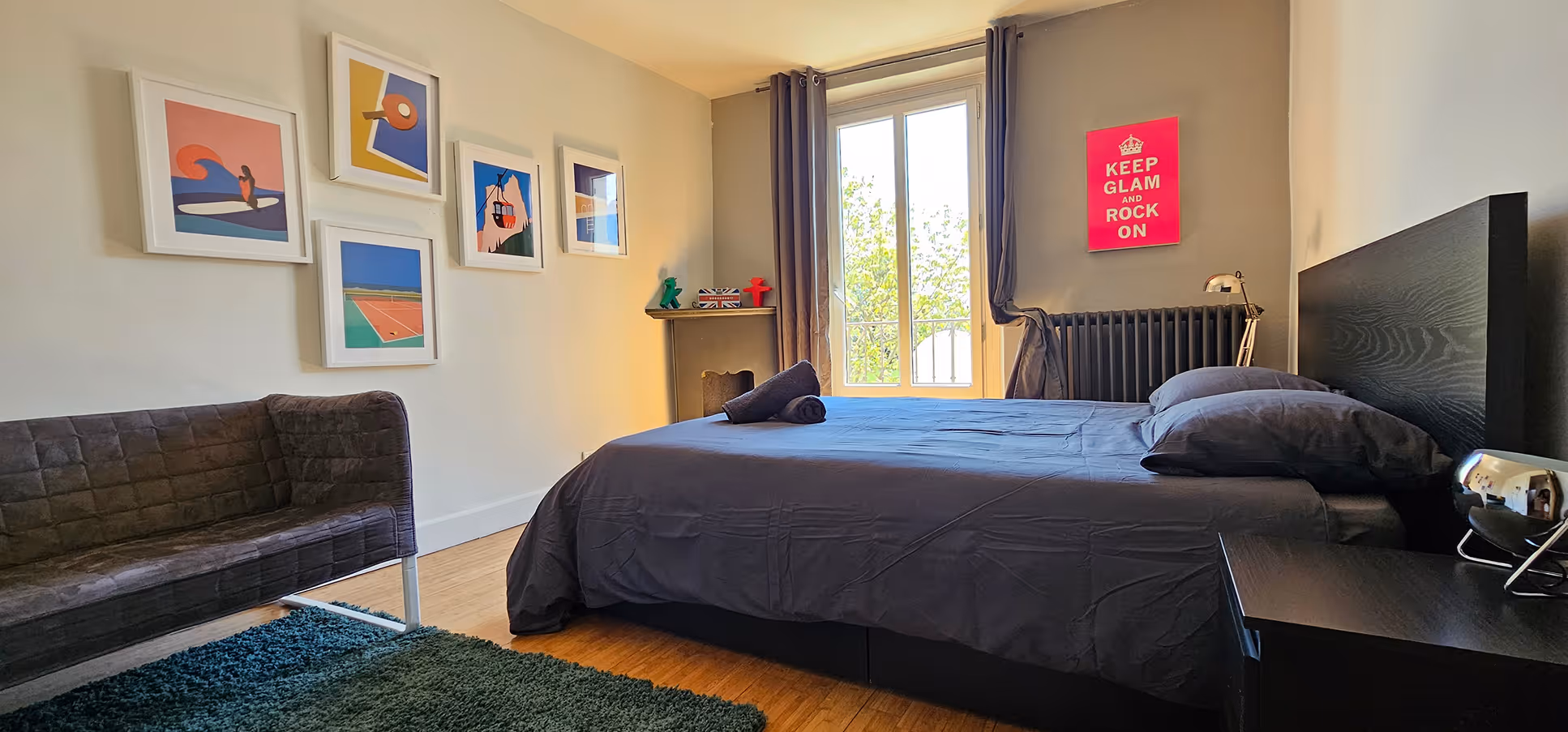 Bright room with a double bed, sofa, colorful paintings, and a red 'Keep Glam and Rock On' poster.