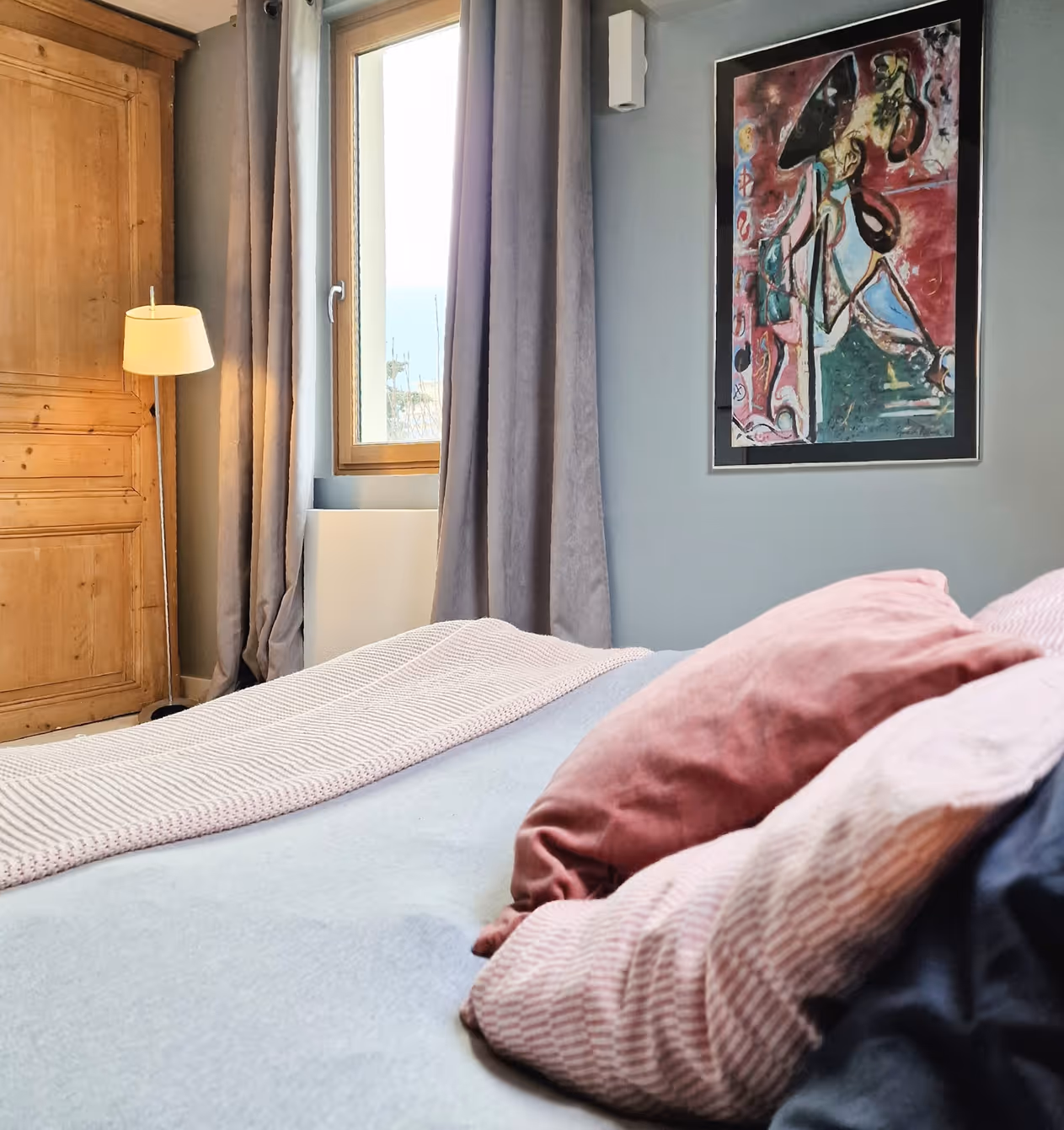 Room with a bed, pink cushions, a large wooden wardrobe, a floor lamp, and an abstract painting on the wall.