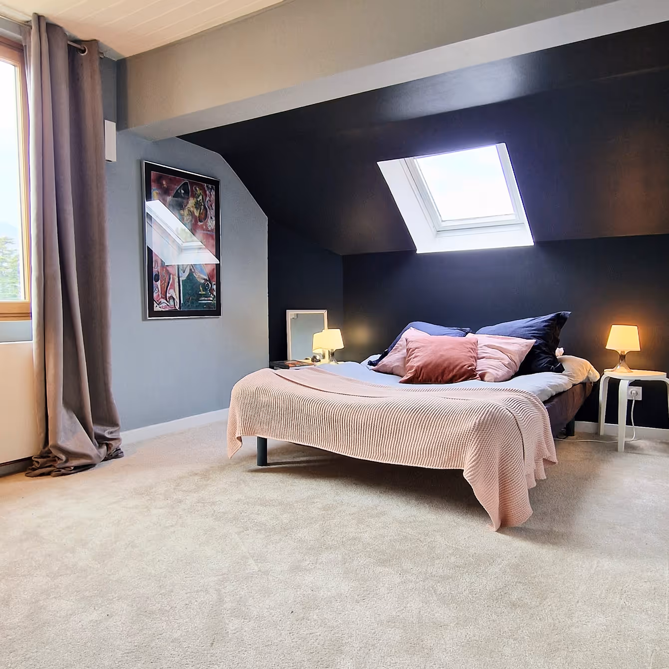 Room with a double bed, a pink blanket, two lamps, a dark wall, and a roof window.