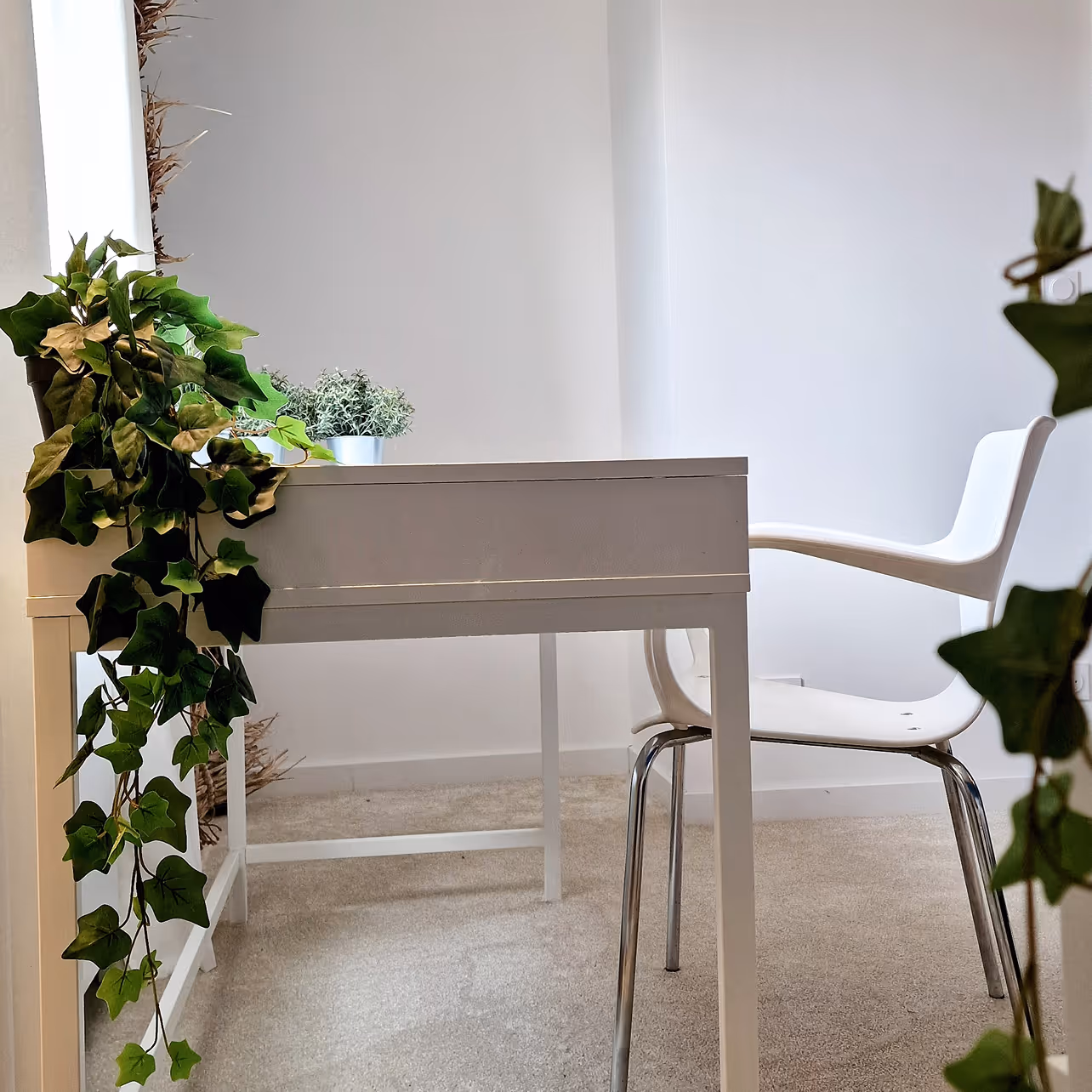Bureau blanc simple avec plante en pot et chaise blanche sur moquette beige dans une pièce lumineuse.