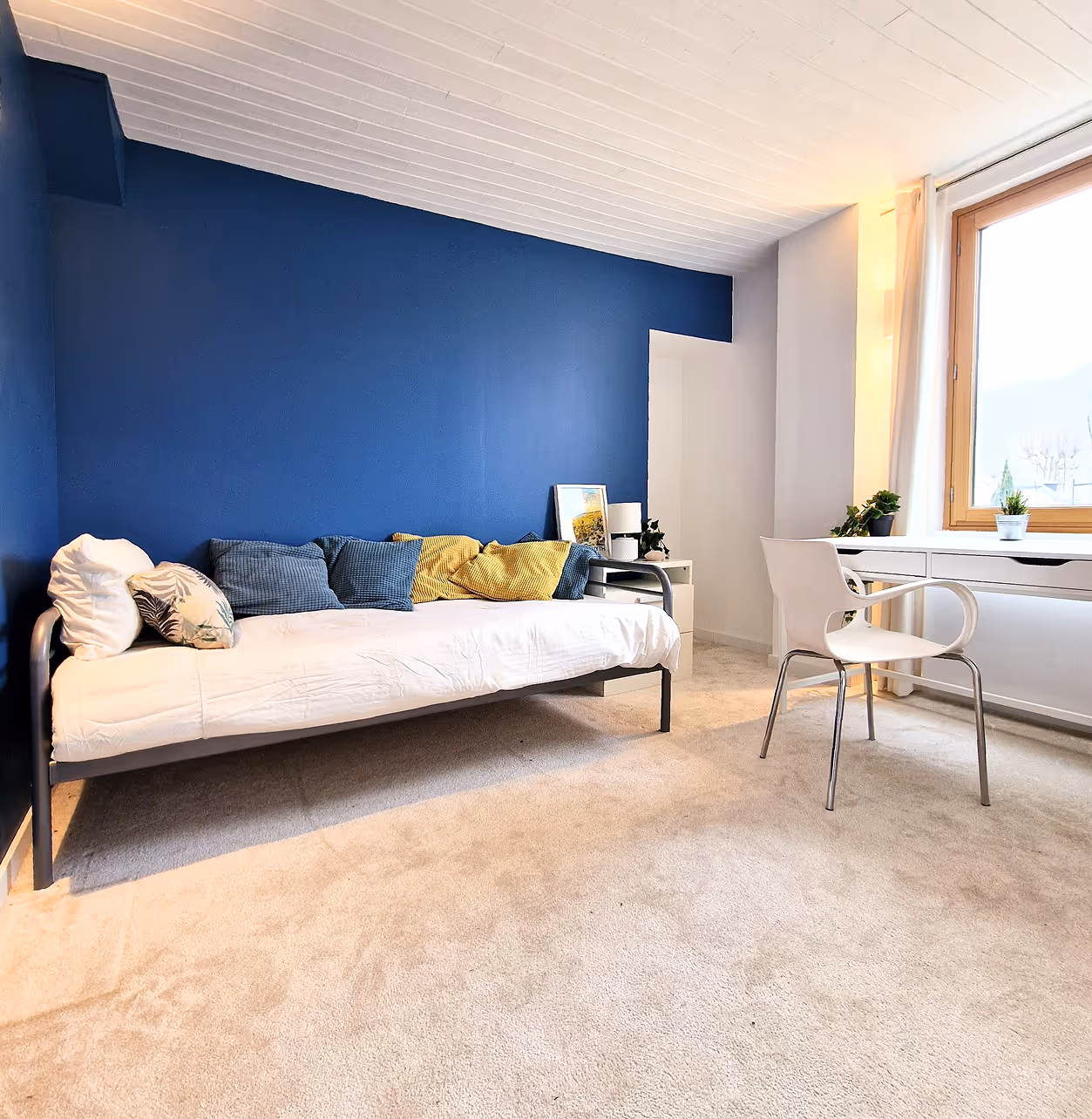 Chambre avec canapé-lit blanc, coussins colorés, mur bleu, chaise blanche et bureau près d'une fenêtre.