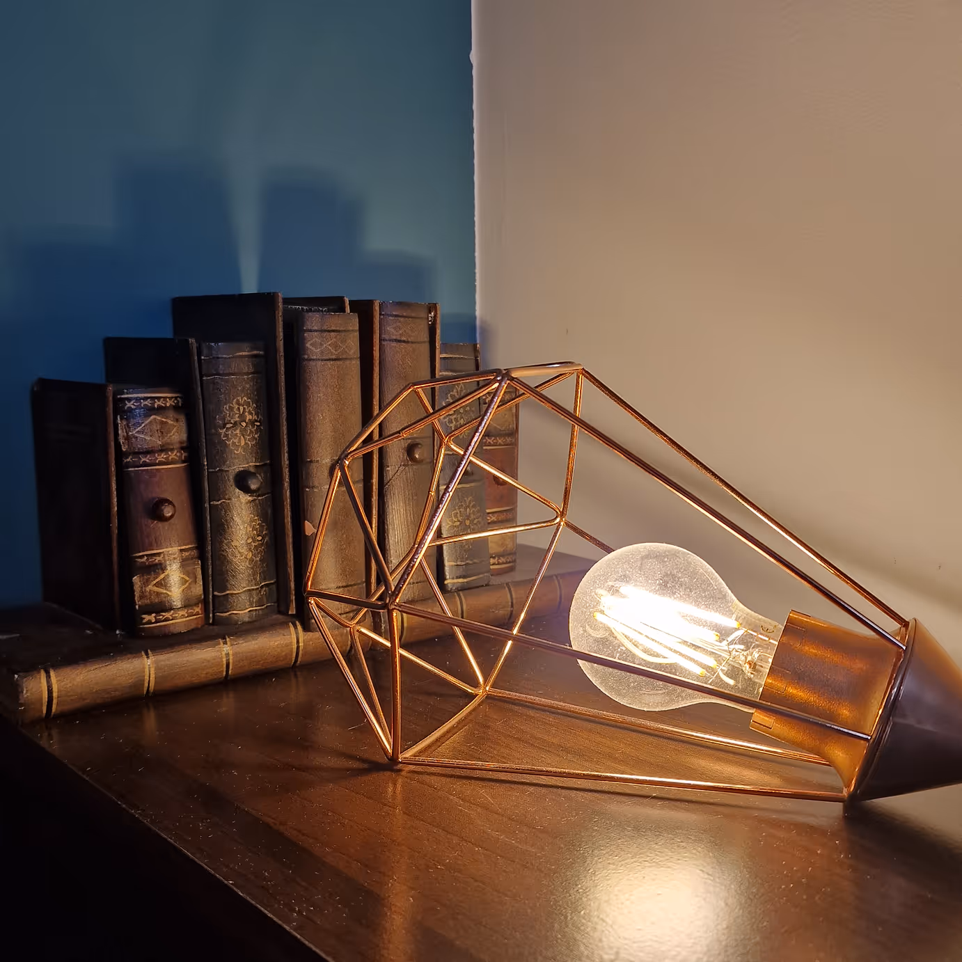 Lampe à ampoule LED dans une structure métallique cuivrée à côté de livres anciens sur une table en bois.
