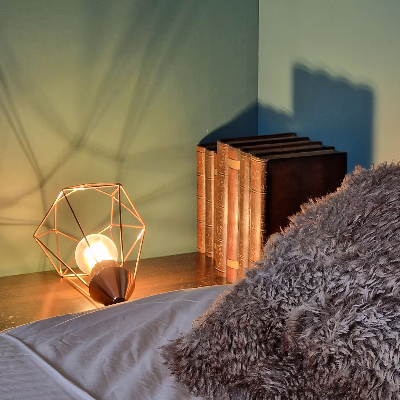 Lampe stylisée sur table à côté de livres anciens et d’un coussin pelucheux gris.