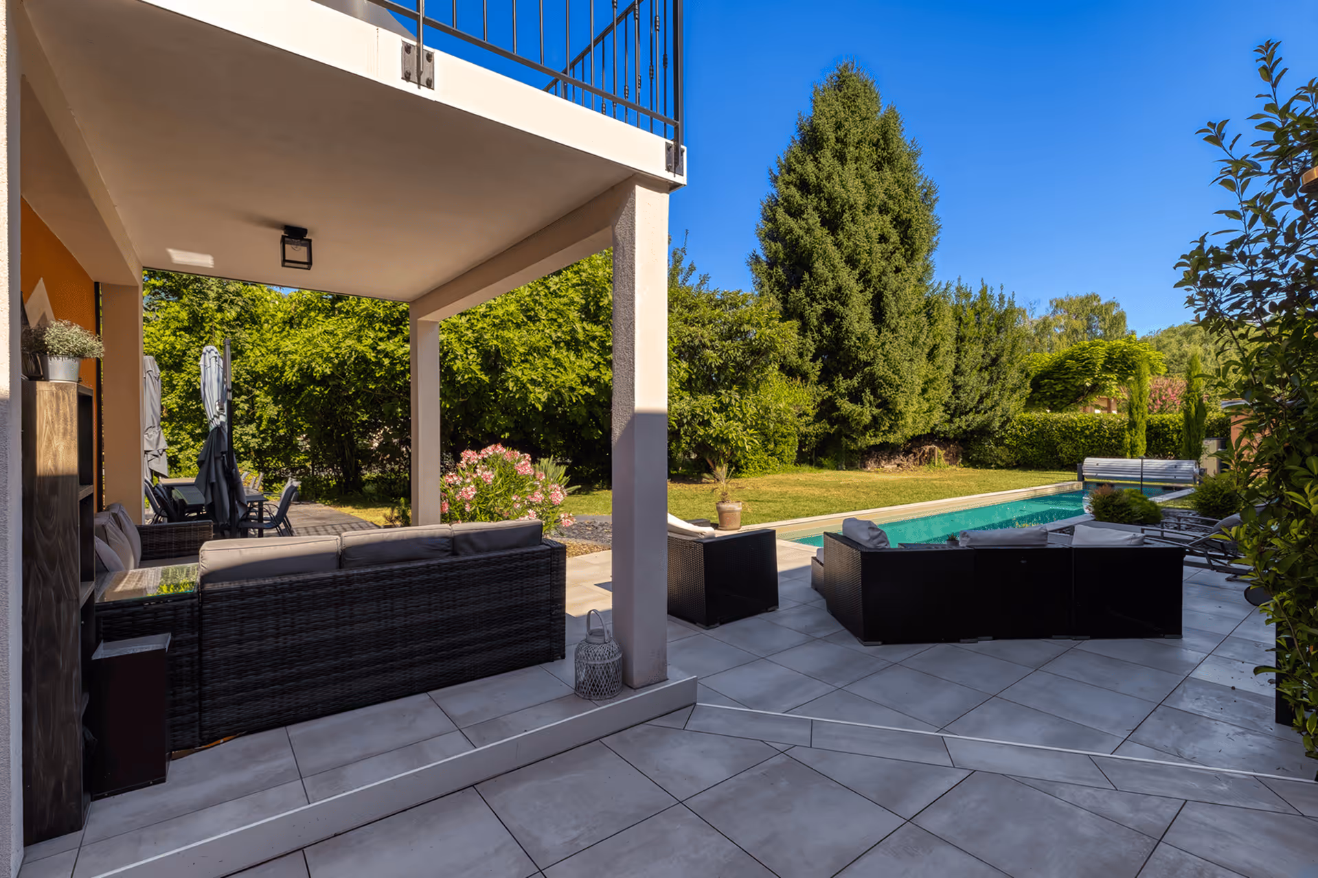 Terrace with rattan sofas, coffee table, red rug, and a pool surrounded by greenery in the sun.
