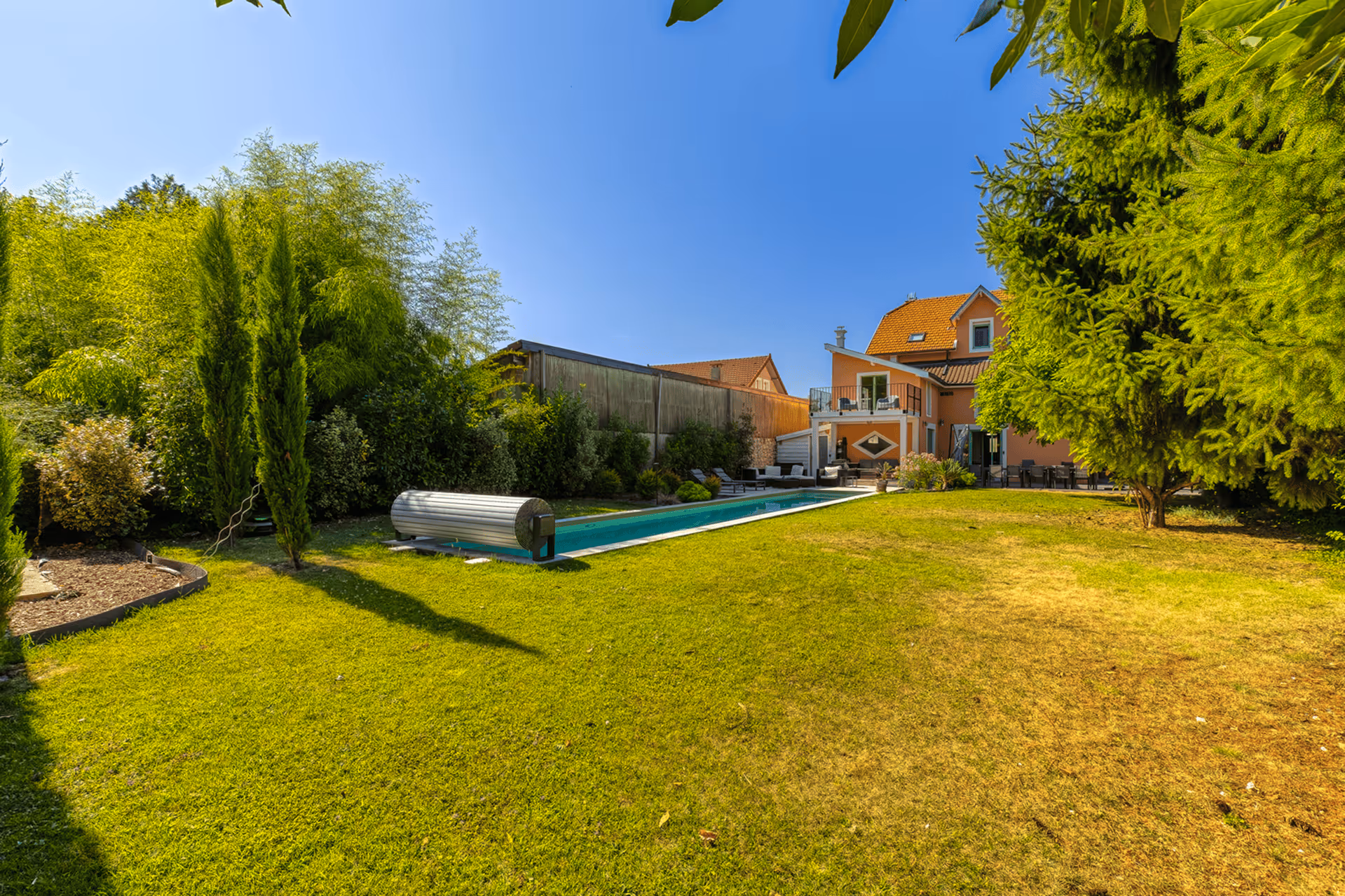 Terrace with rattan sofas, coffee table, red rug, and a pool surrounded by greenery in the sun.