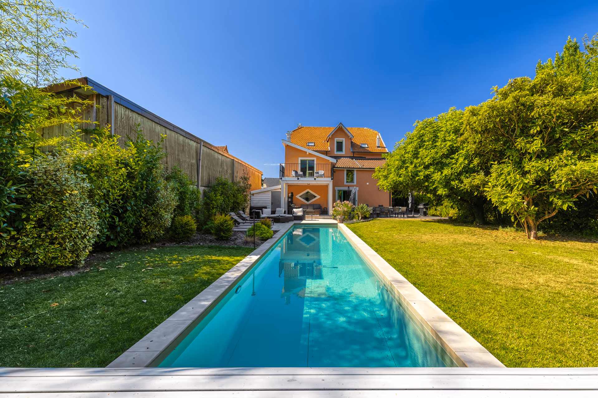 Rectangular pool surrounded by sun loungers and armchairs, a lush garden, and a clear blue sky.