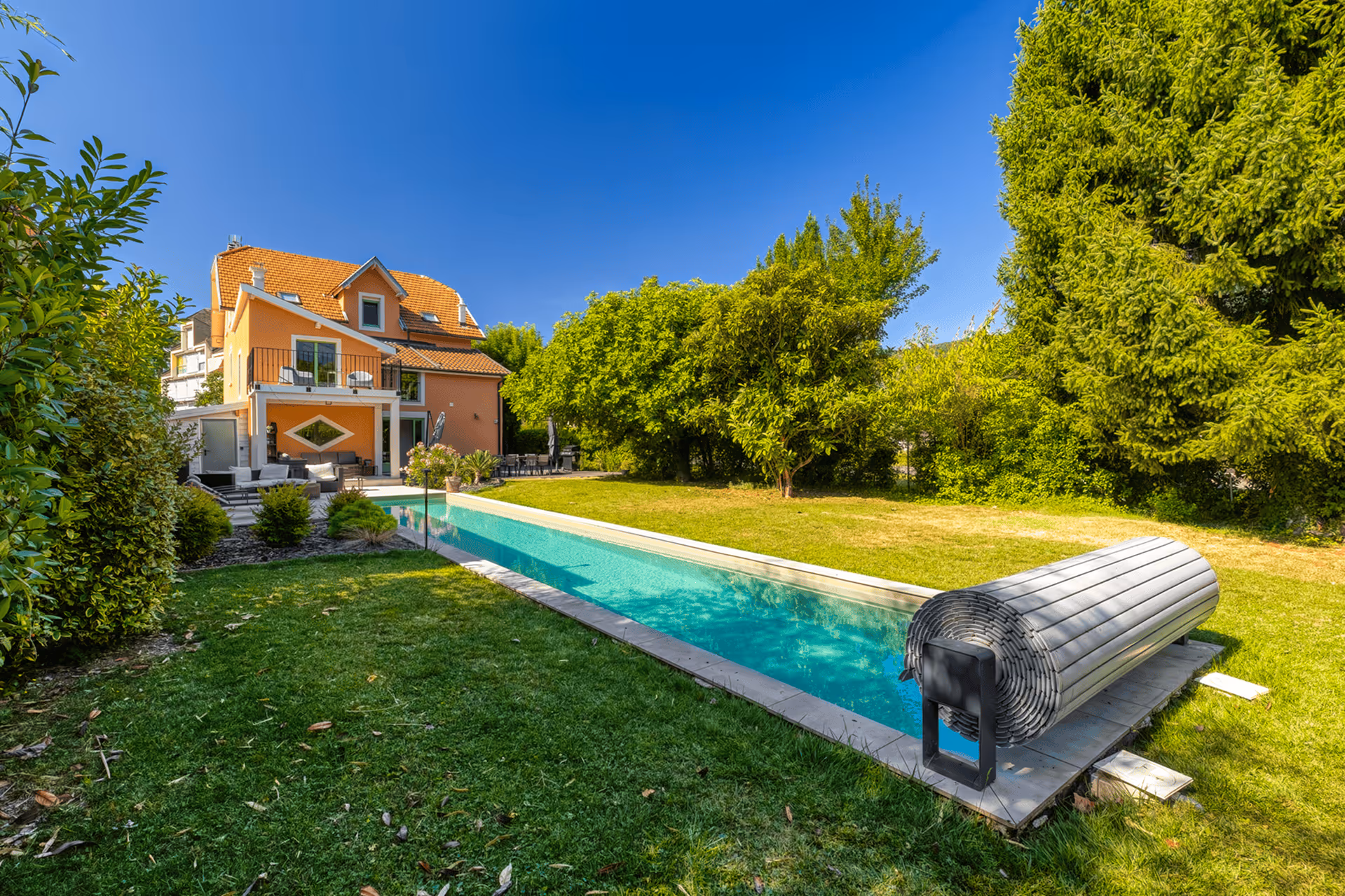 House with a long pool and a lush garden under a clear blue sky.