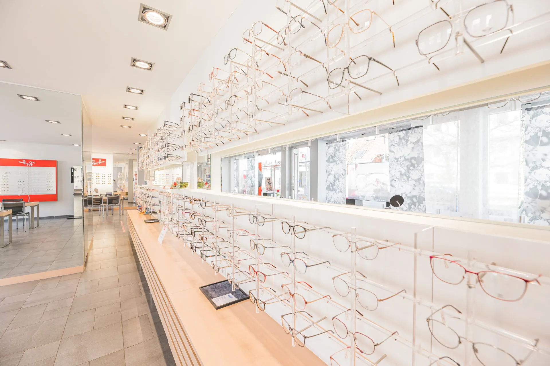 Helle Optikerboutique mit Regalen voller Brillenfassungen entlang der Wand und Sitzgelegenheiten im Hintergrund.