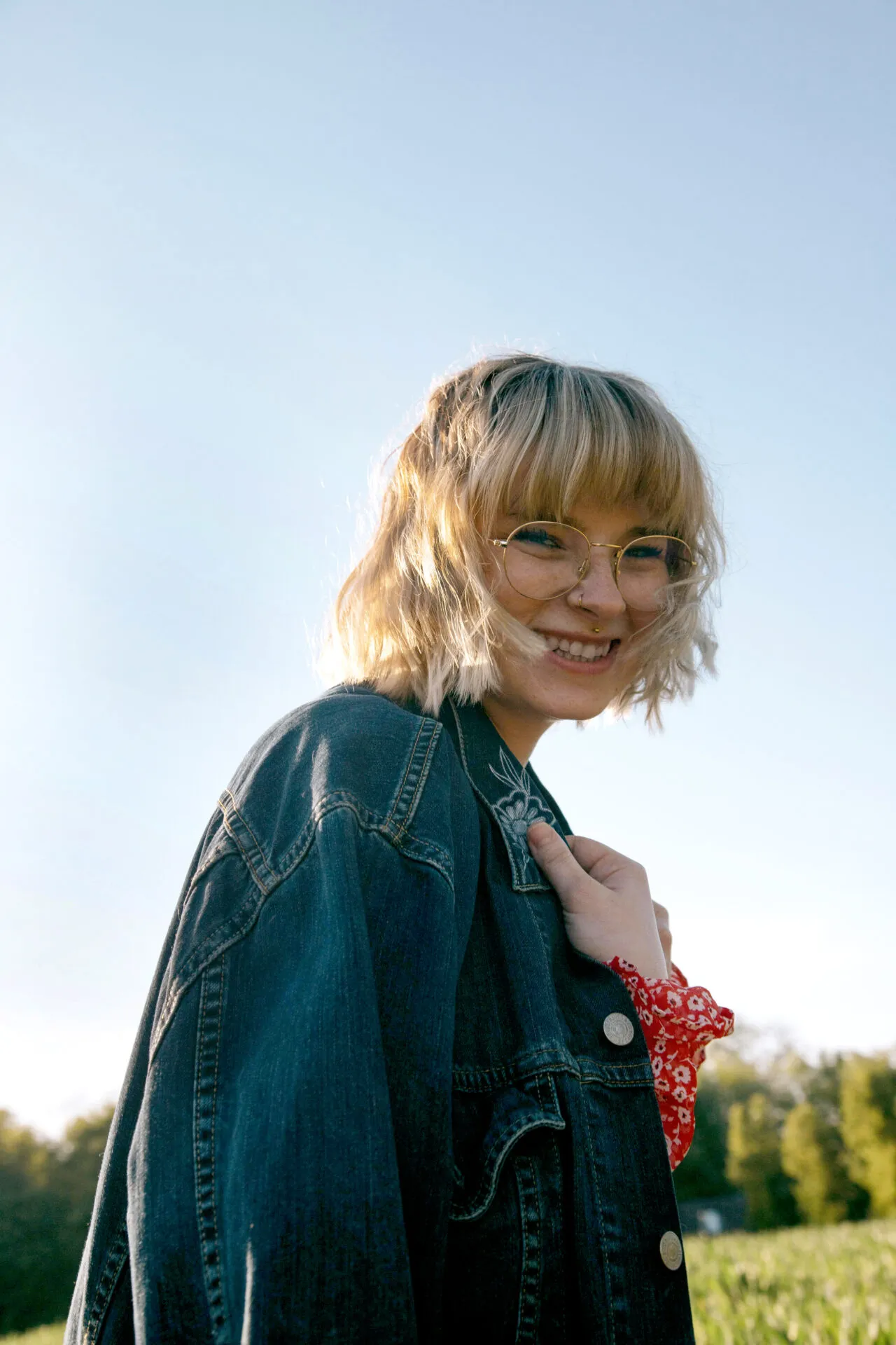Junge Frau mit blonden Haaren, Brille und Jeansjacke lächelt draußen im Sonnenlicht.