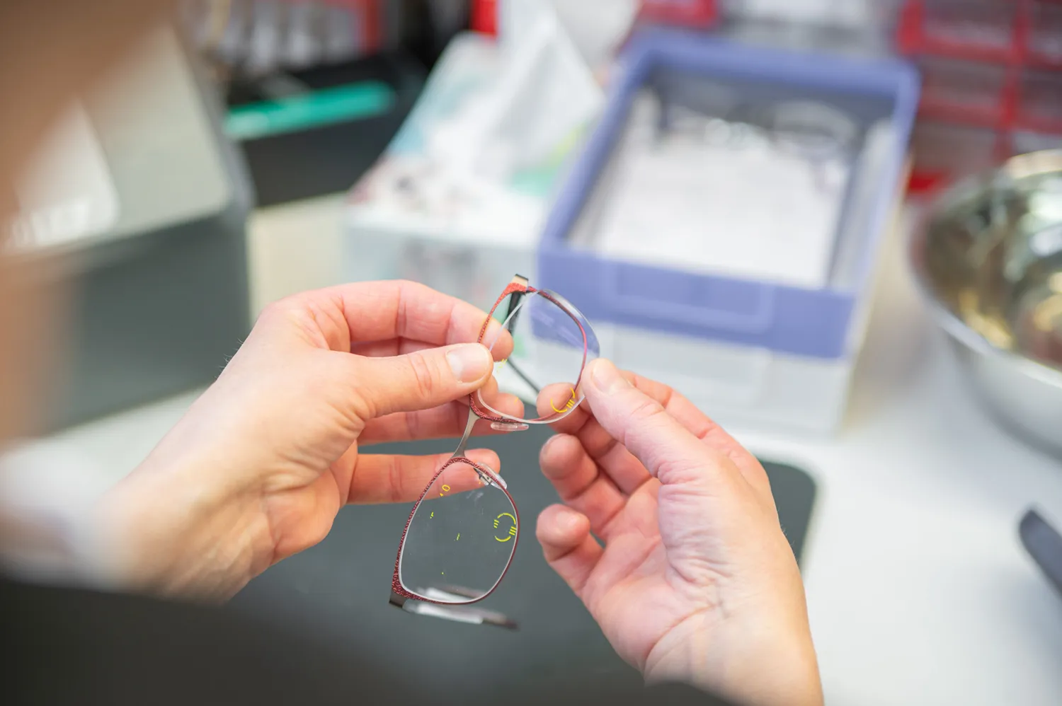 Jemand hält eine Brille mit roten Rahmen vor unscheinbarem Werkstatt-Hintergrund.
