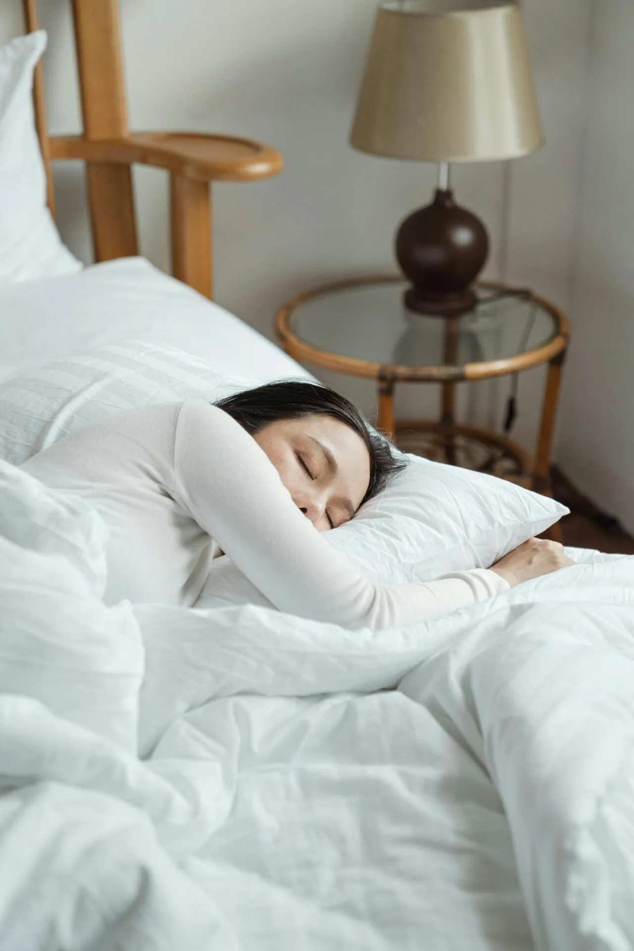 Frau schläft auf dem Bett mit weißer Bettwäsche und einem Kissen, neben einem Nachttisch mit Lampe.